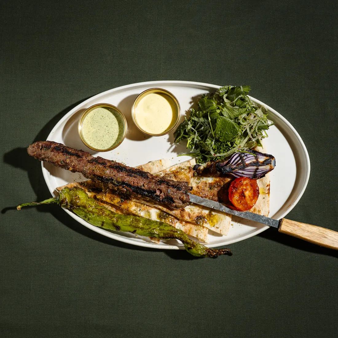Kofta kebab on a skewer over fresh laffa with grilled vegetables and sauces