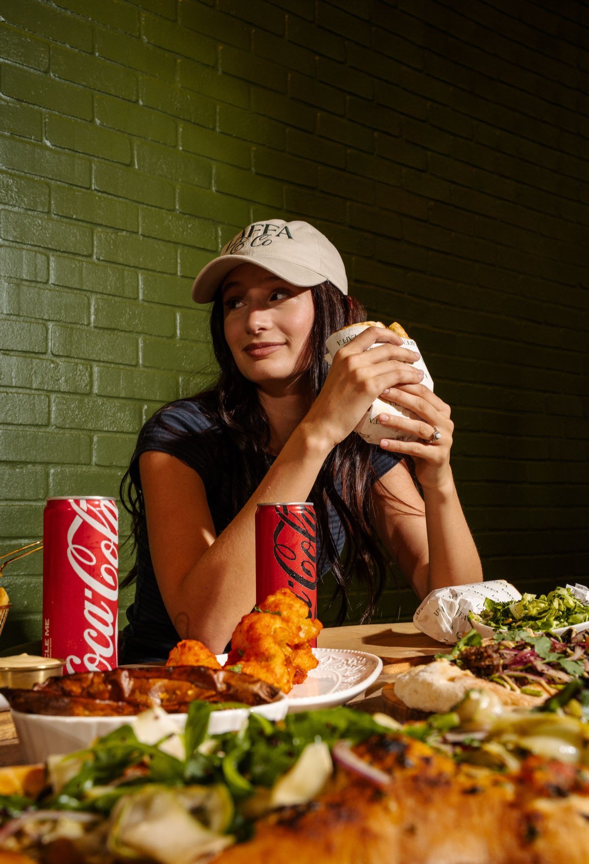 Guest in a Laffa & Co cap holding a wrap at the olive-green brick wall